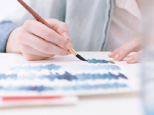 close up image of a person in a light blue denim shirt painting blue watercolor stripes on white paper.  