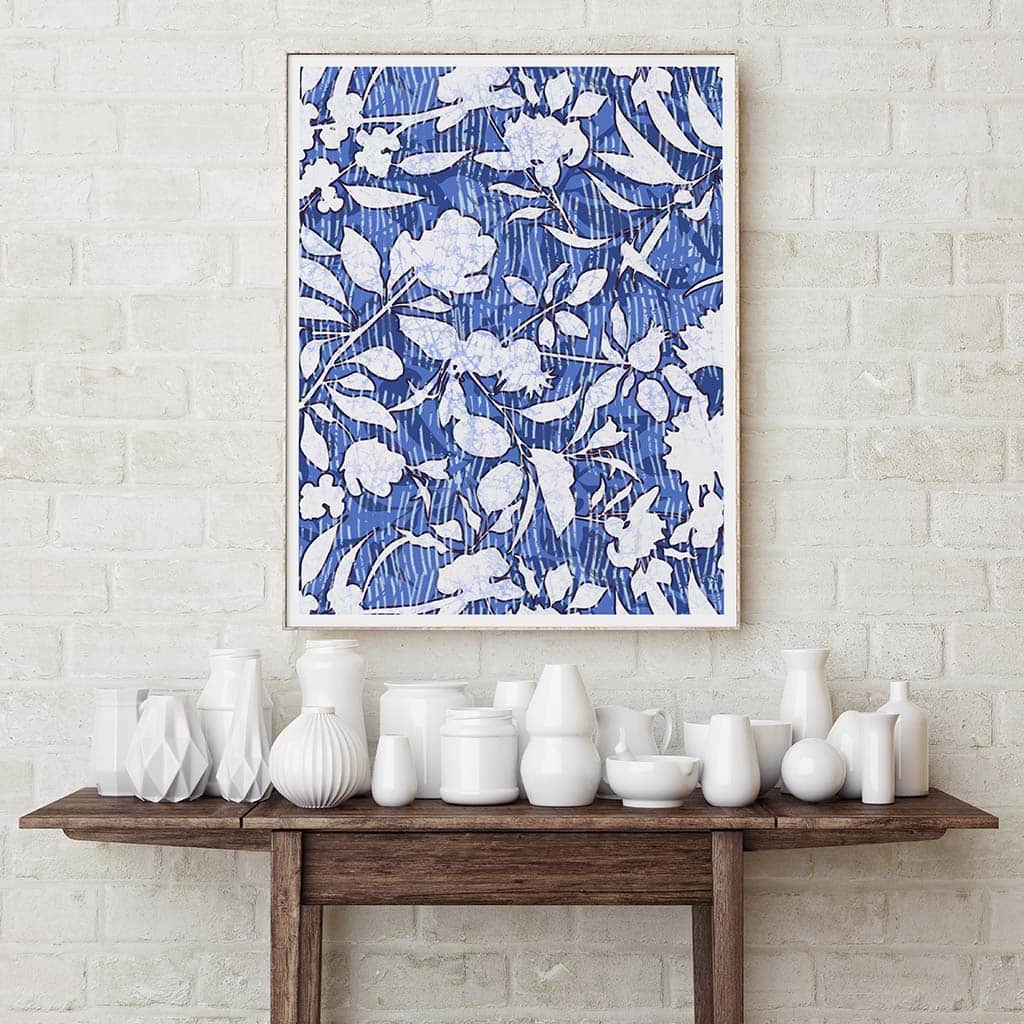 Blue &amp; White Fine art print by Jenny Bova. Blue linear pattern in background with white silhouetted floral and leaf shapes in the foreground. Small white border for artists signature. Print is on a white brick wall over a wood table filled with white porcelain vases