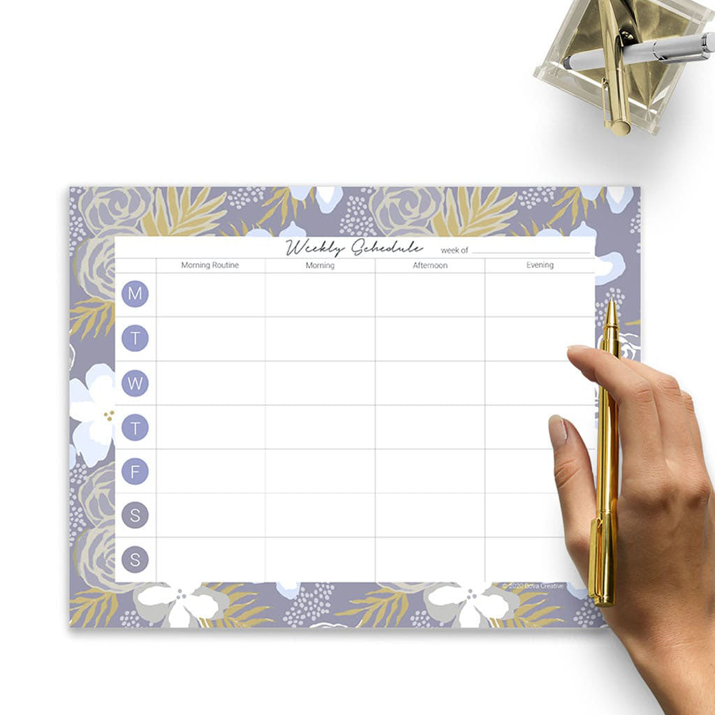 Weekly Planning Pad with floral botanical border pattern on a white surface. Woman's hand holding gold pen, pencil cup in upper right corner