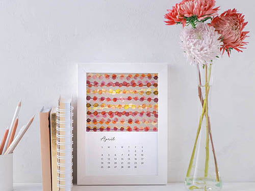 april month of jenny bovas 2021 desk calendar in a white frame on a desk, next to flowers and office supplies