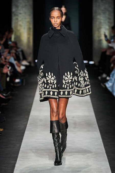 Runway image from A/W 2020 Ermanno Scervino fashion show. Woman wearing tall black boots and a black cape coat embroidered with white florals