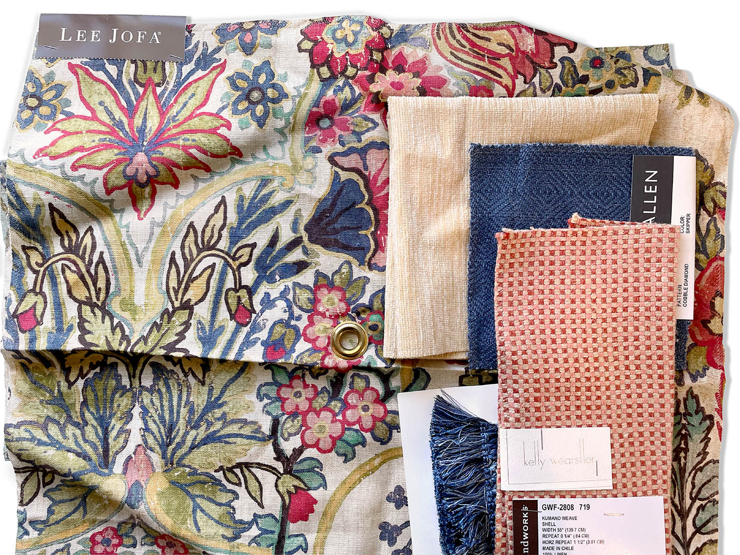 Interior design materials are layered on top of one another. Lee Jofa printed fabric with blue, yellow, red, and green botanicals. Solid off white chenille, solid blue twill, pink and red check, and blue fringe trim.