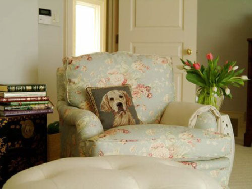 image of an upholstered chair with a golden retriever pillow on it. The chair has a floral pattern in light blue. In the foreground is a cream colored ottoman. On the left is a small chest stacked with books. On the right is a vase of tulips. 