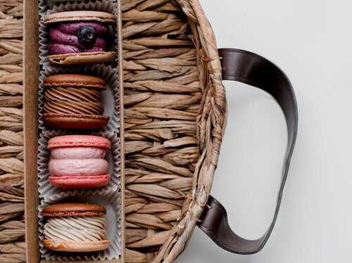 image of colorful macarons in a box. the box is on a basket/tray with leather handles. The background is white. 