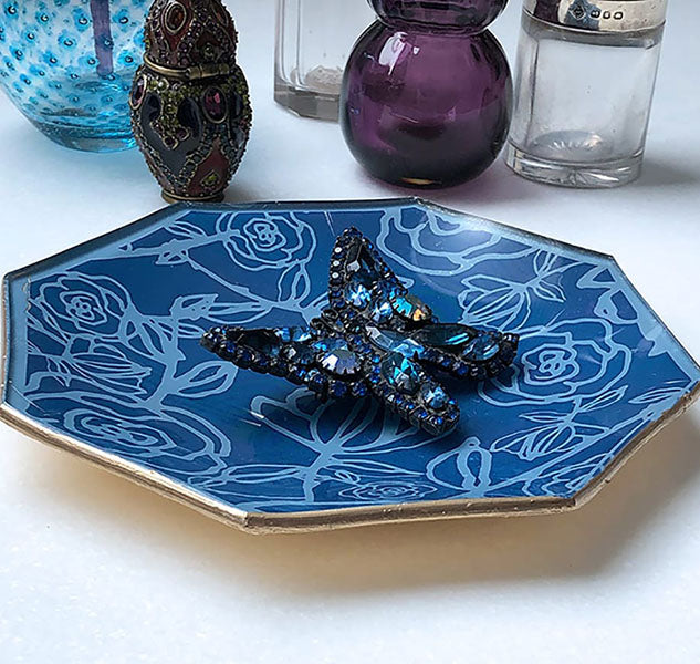 Glass jewelry tray with blue graphic pattern and gold edging on white surface