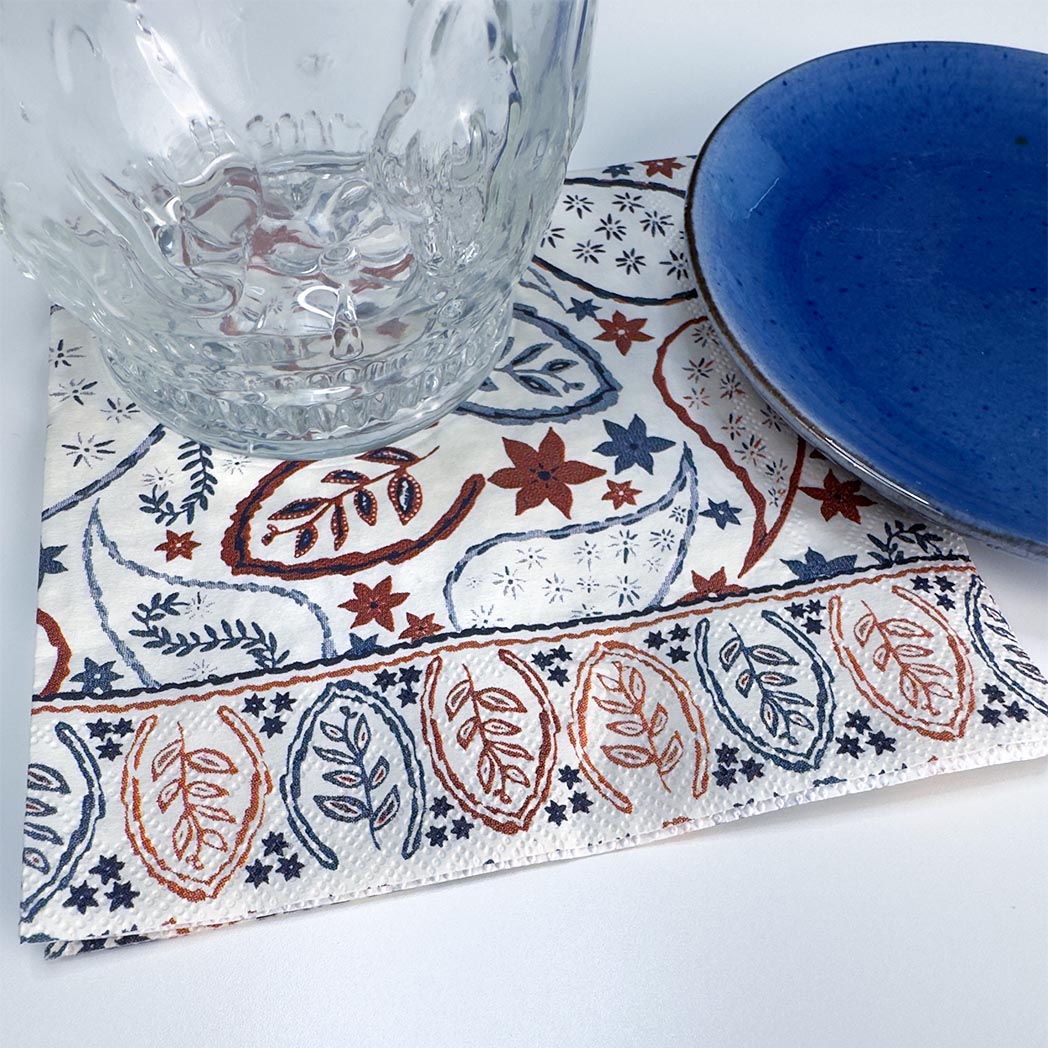A square printed placemat with a white base and blue and rust-red paisley and floral motifs, styled with a clear glass tumbler and a blue ceramic plate on a white surface.