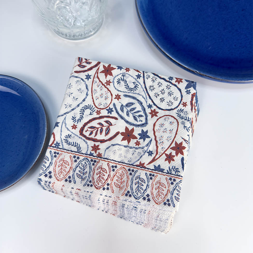 A folded white fabric napkin with a red and blue paisley and floral block print pattern, styled on a white surface with blue ceramic plates and a glass.