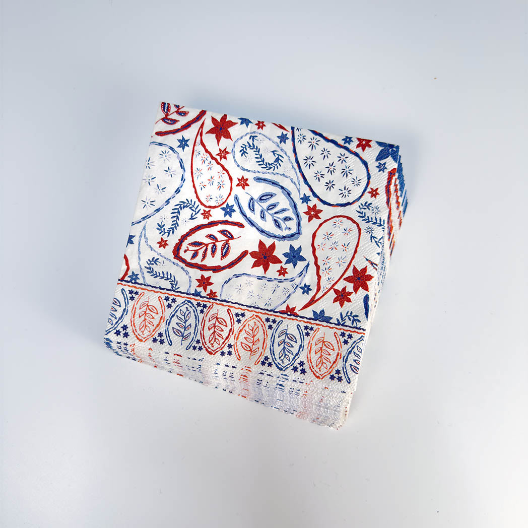 A square white fabric napkin or table linen with a red and blue paisley block-print pattern and a decorative border, shown on a light gray background.