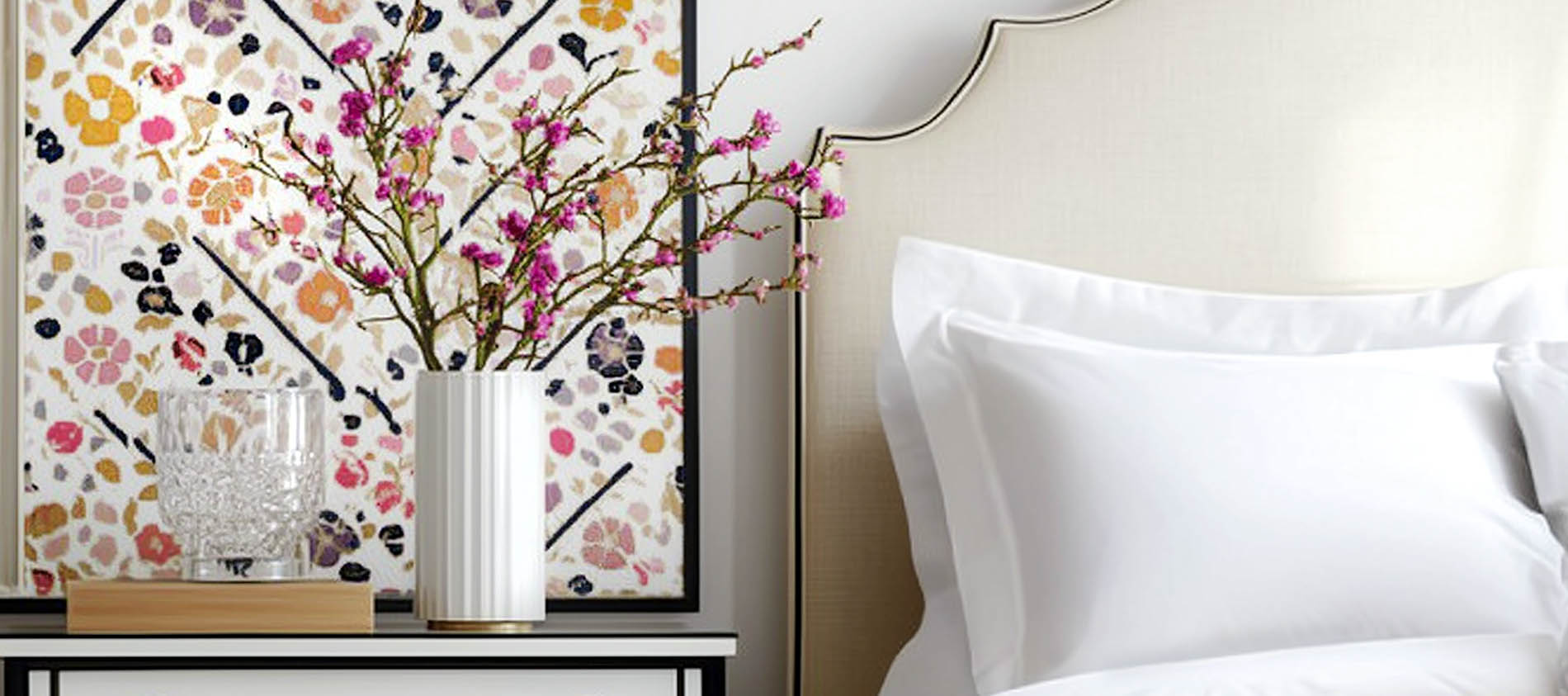 crisp headboard and bedding with bright floral artwork and vase of pink flowers