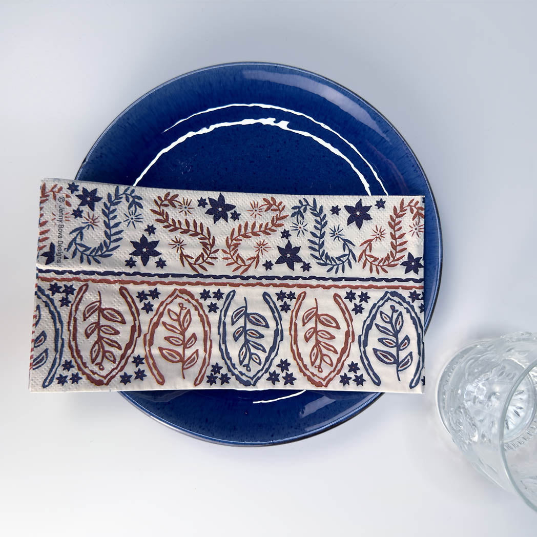 A rectangular cream fabric napkin with a detailed navy and rust-red botanical block print pattern, folded and displayed on a glossy dark blue ceramic plate, with a clear glass partially visible on the right.
