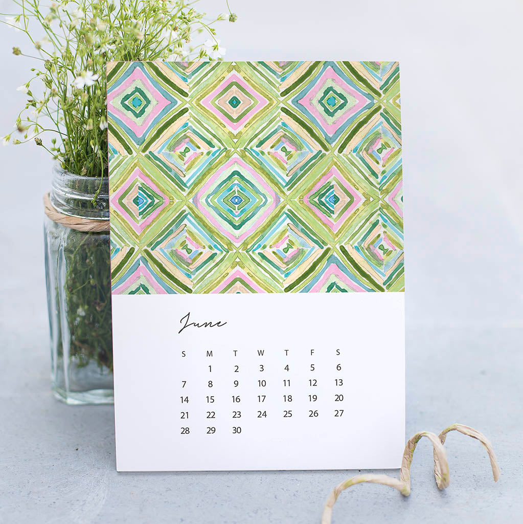 Calendar with geometric pattern and June month displayed, placed next to a vase with greenery on a light surface.