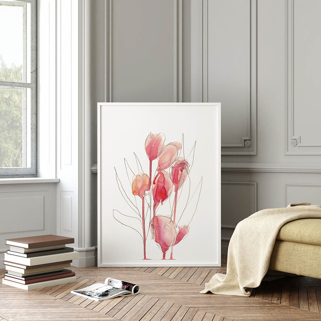 24x36 Fine art print by Jenny Bova shown in a white frame leaning against a gray wall in a living room.  The print features an abstract tulip bouquet pencil sketch partially filled in with loose pink watercolor. Small white border for artists signature. Books are stacked to the left of the print, golden yellow upholstered furniture is to the right.