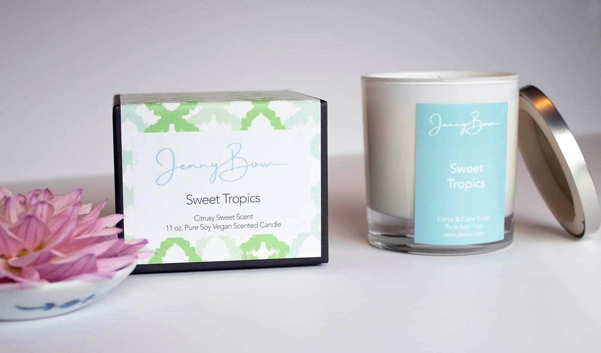Citrus scented soy candle with a light blue label and silver lid leaning against the side. Black candle box with patterned label and a pink flower sit next to the candle. 