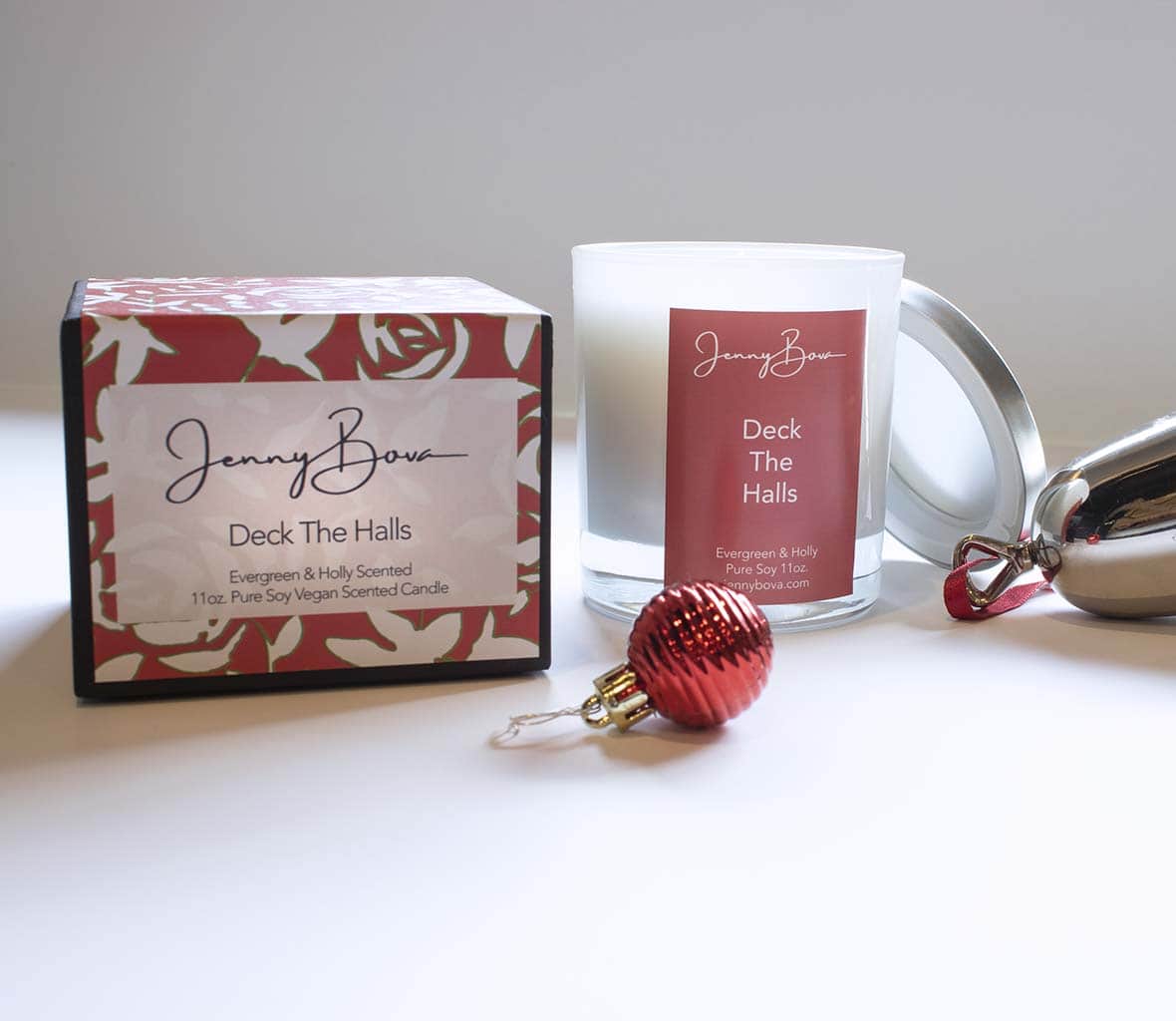Evergreen scented soy candle with a red label next to a box with a patterned label. In front of the candle is a mini red christmas ornament and off to the side is a silver bell with red ribbon. 
