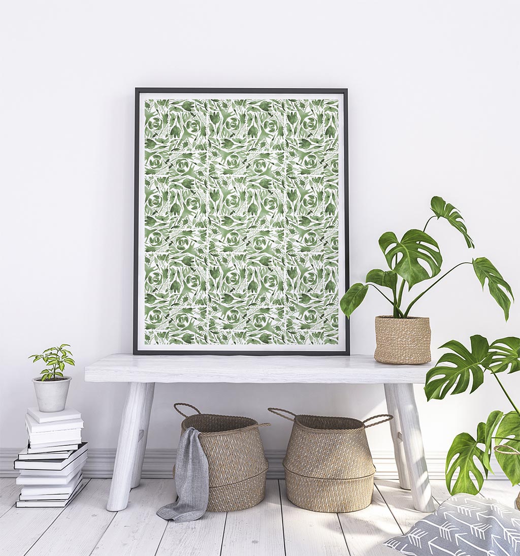 Repeating watercolor floral design in sage greens in a large black frame which on top of a white bench leaning on the wall. Around bench are plants, baskets, and books