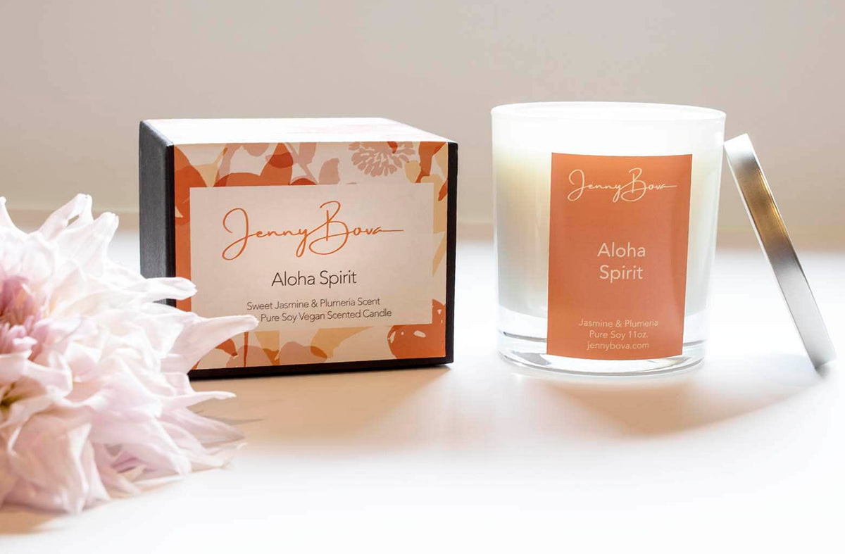 Jasmine &amp; Plumeria scented soy candle with orange label, black box with patterned label, and a dahlia on a white background