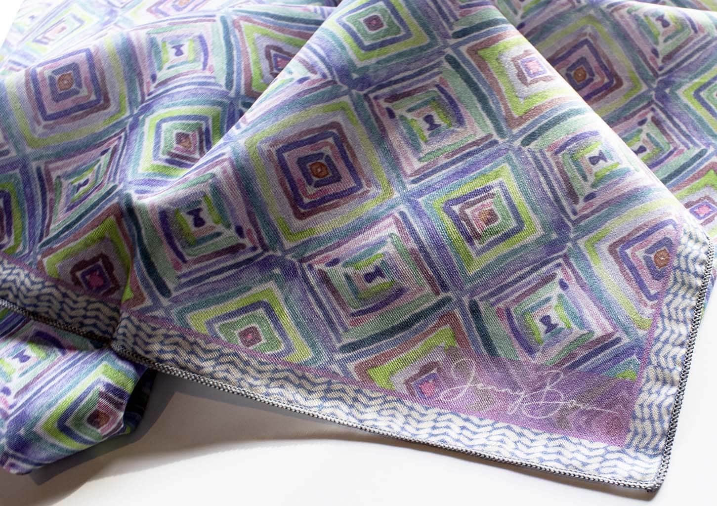 corner detail of a silk scarf with a blue, green, and purple watercolor geometric design. The Jenny Bova logo is printed on the corner shown.