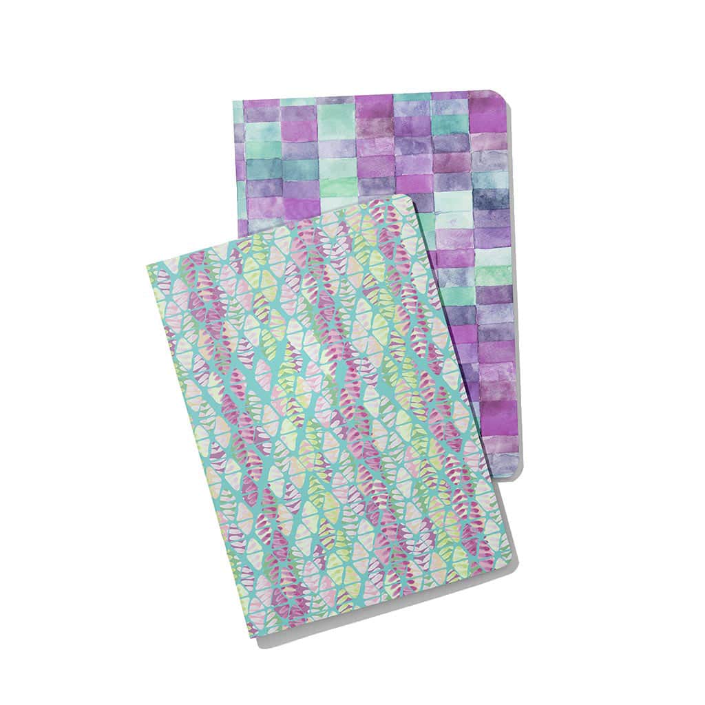 2 soft cover notebooks with teal, lavender, purple, aqua, and pink designs. The one on the top is a triangular design, and the one behind is watercolor rectangles. 