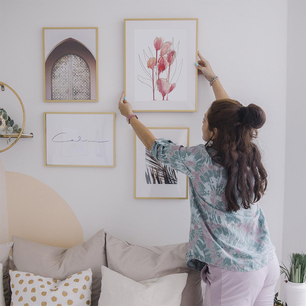 Woman hanging pink watercolor tulip sketch art print on a white wall with other creative artistic prints. Plants, a headboard, and pillows are shown. 